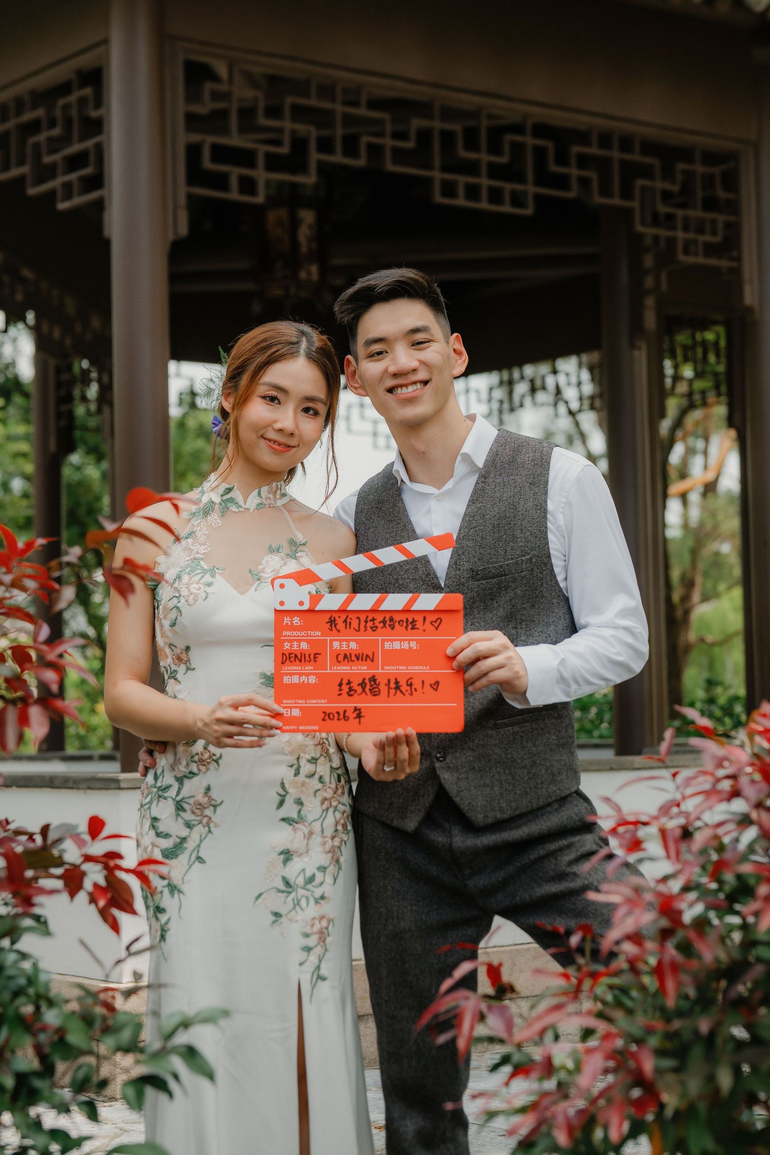 A couple holding red double happiness signs in a traditional Chinese garden setting.