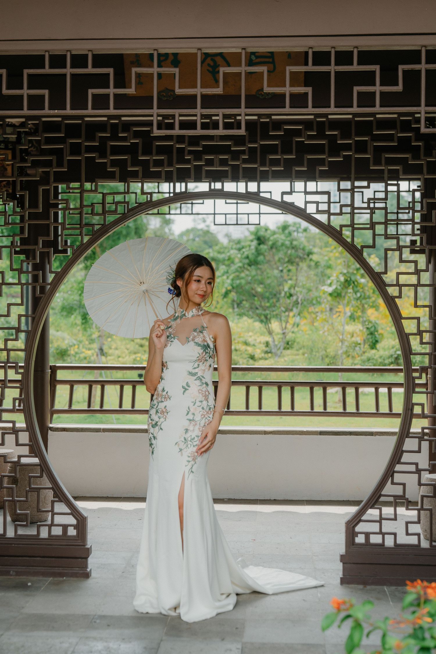 A bride in a flowy white wedding dress poses gracefully within a covered Asian-style garden pavilion setting.