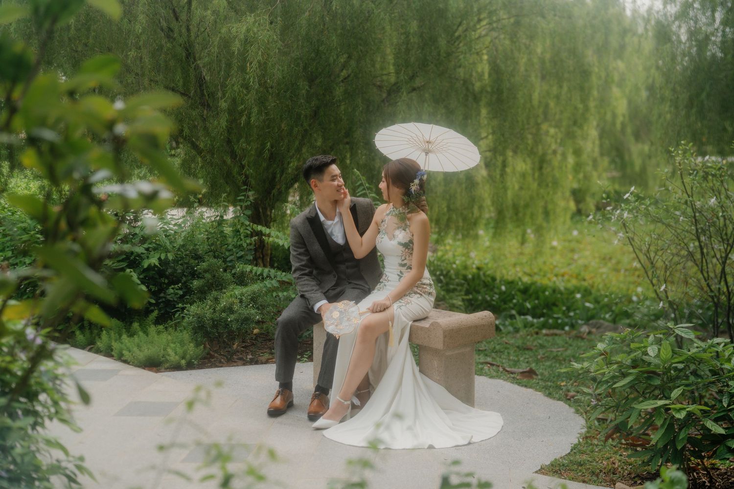 A couple shares an intimate moment on a stone bench under a white parasol in a garden setting.