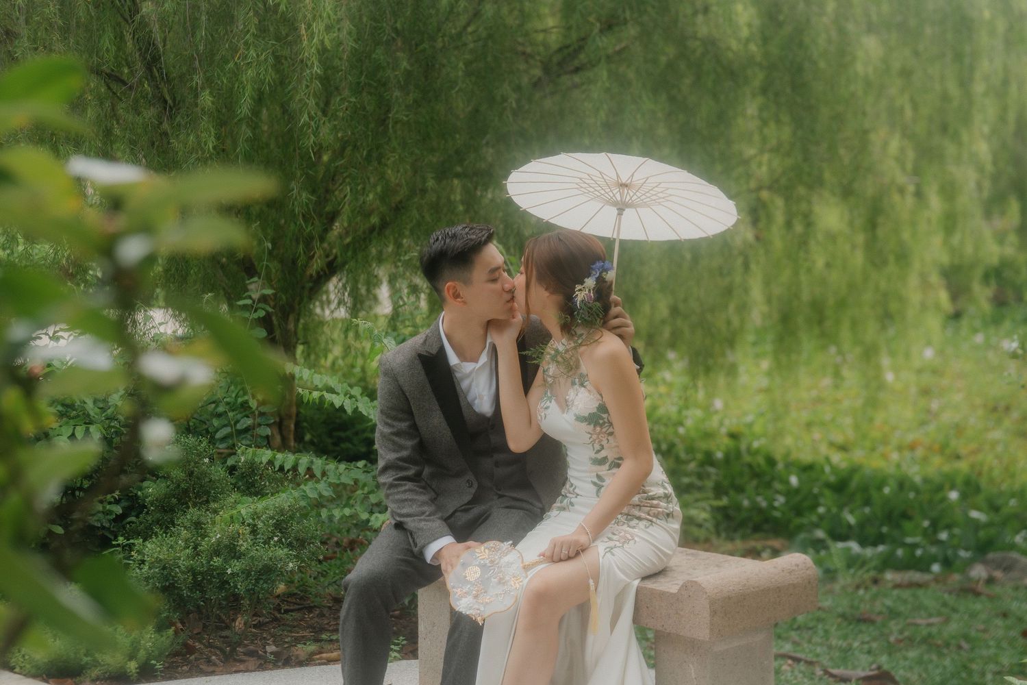 Couple posing with parasol in a serene garden with willow trees.