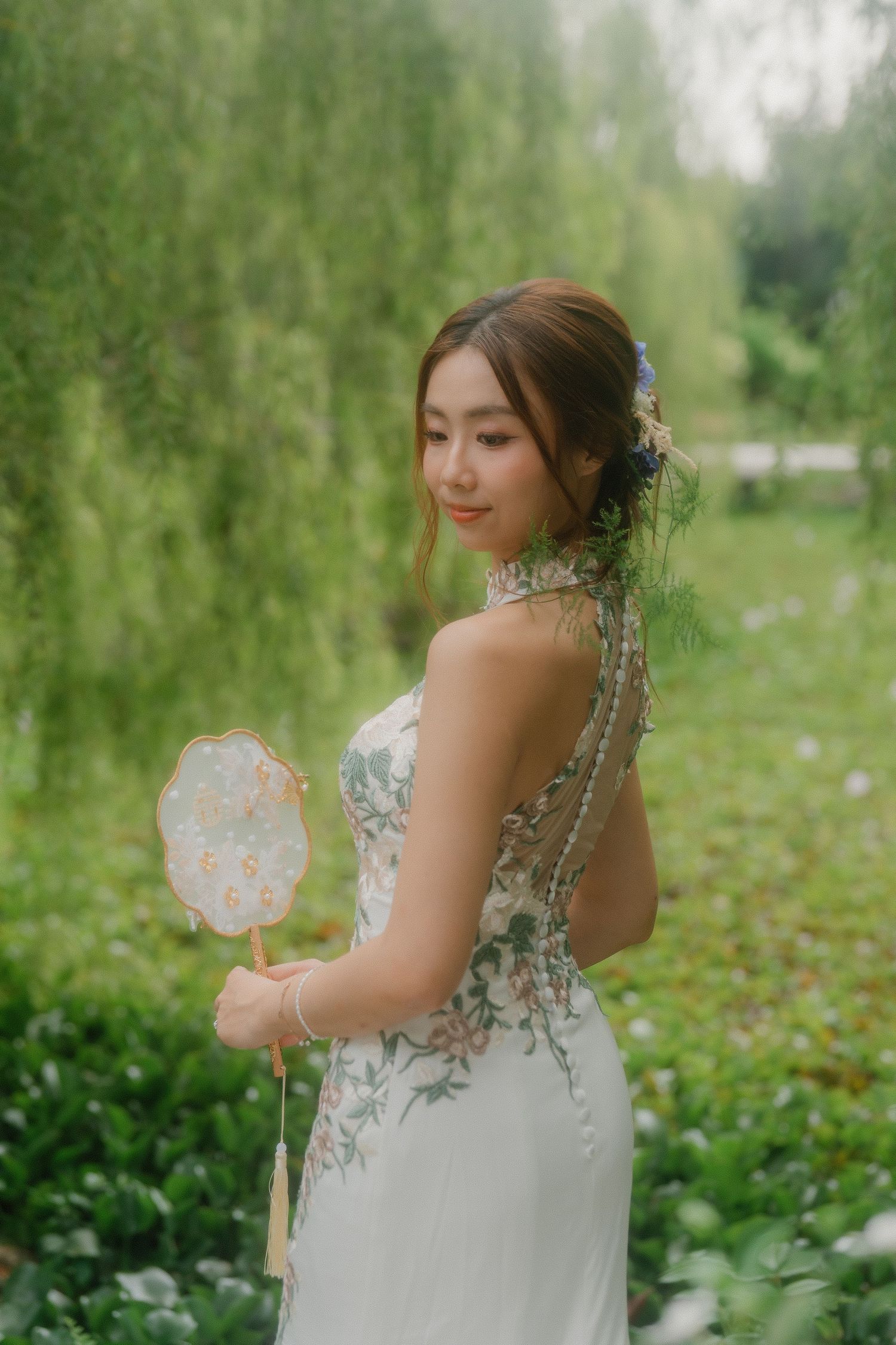 Bride holding decorative fan poses gracefully in garden with floral backdrop.