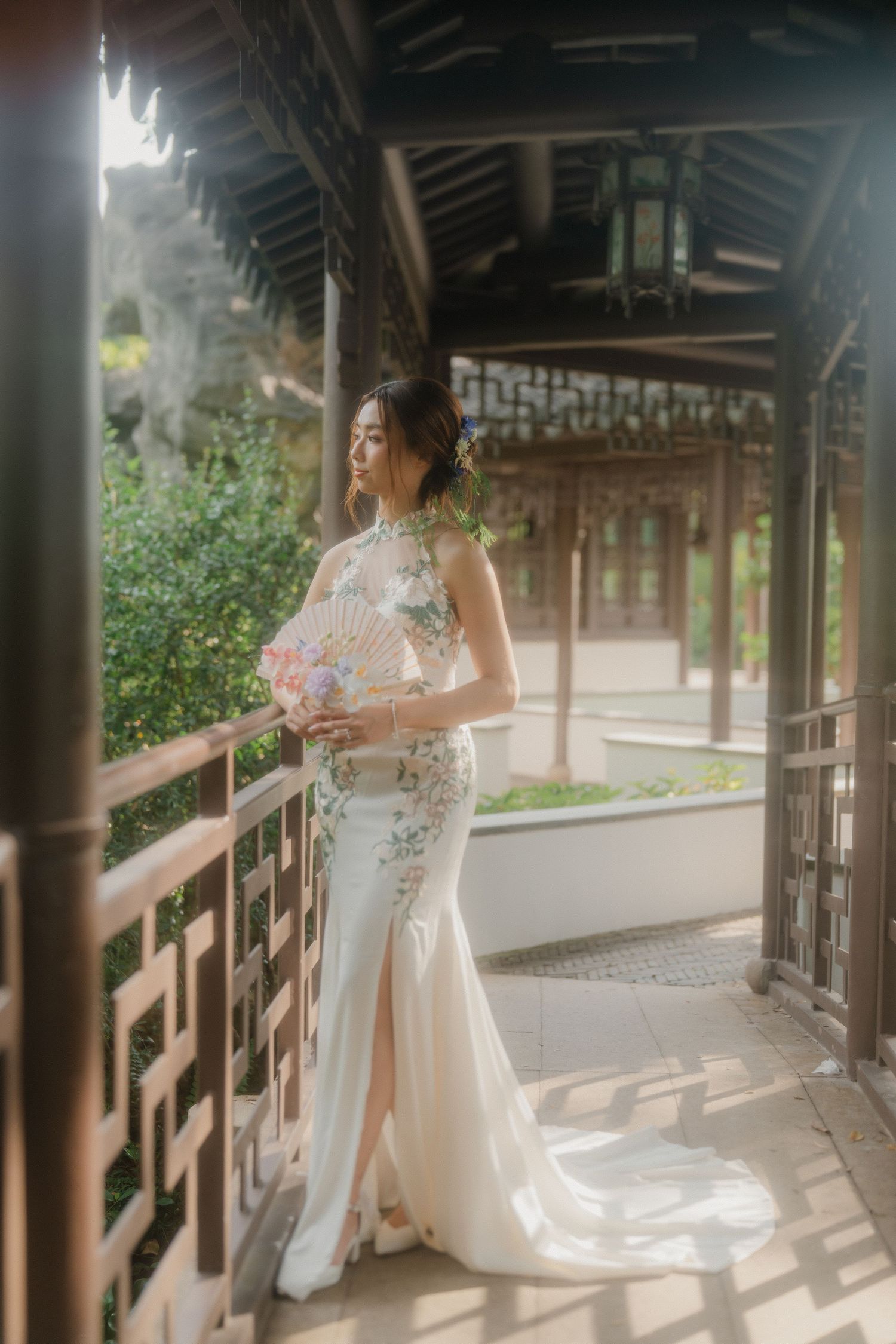 Traditional white qipao gown with floral pattern and train is showcased in covered wooden walkway at a Chinese garden.
