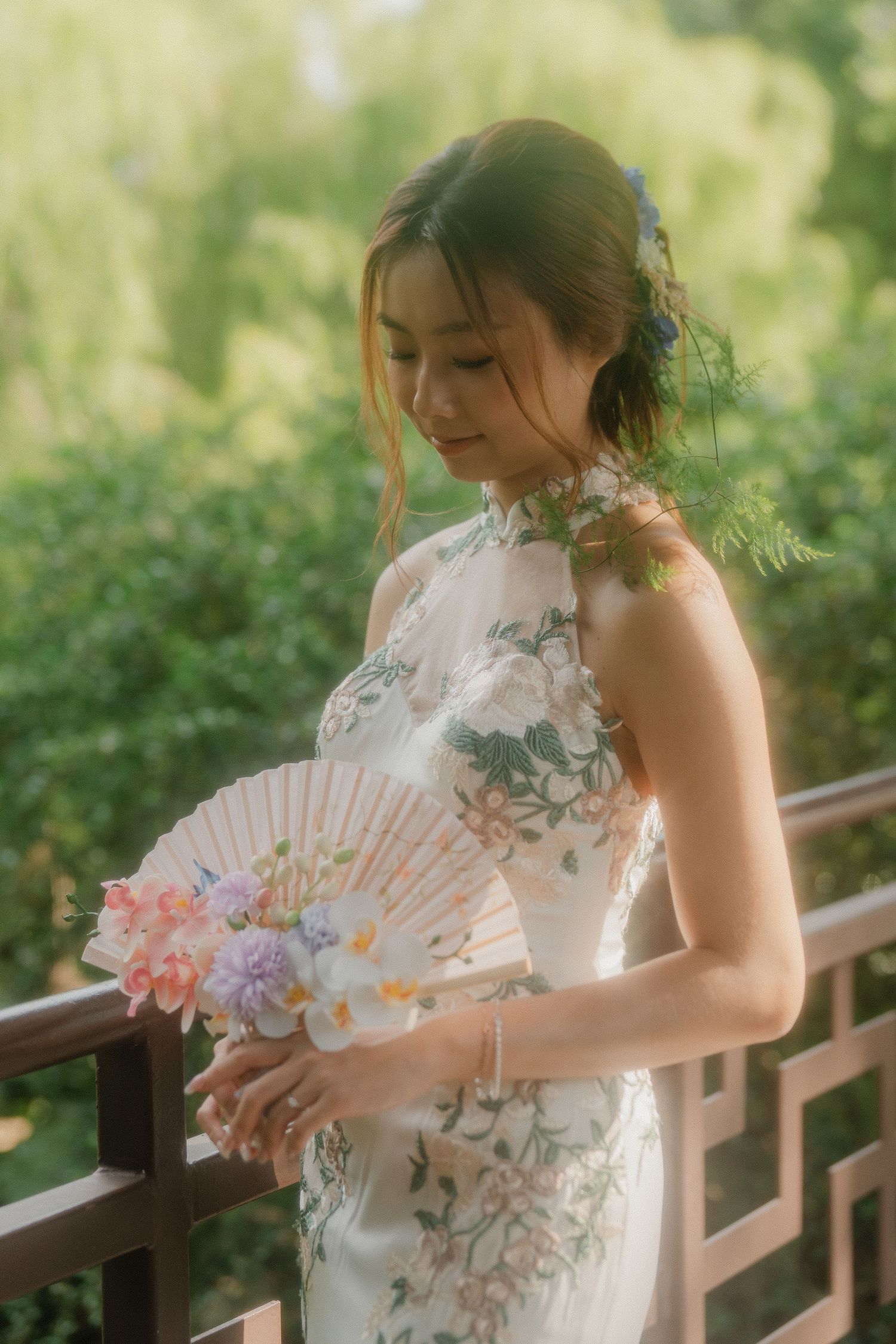 Elegant bride in white cheongsam dress holding a decorative fan while standing on a wooden deck.
