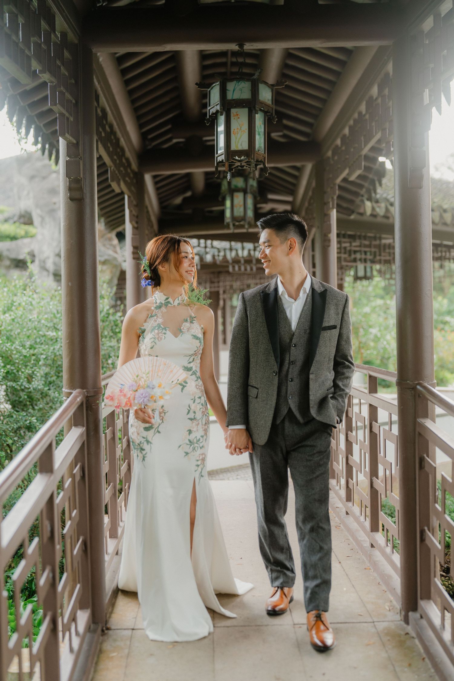 A couple in wedding attire walk and pose together along a covered wooden walkway surrounded by lush greenery.