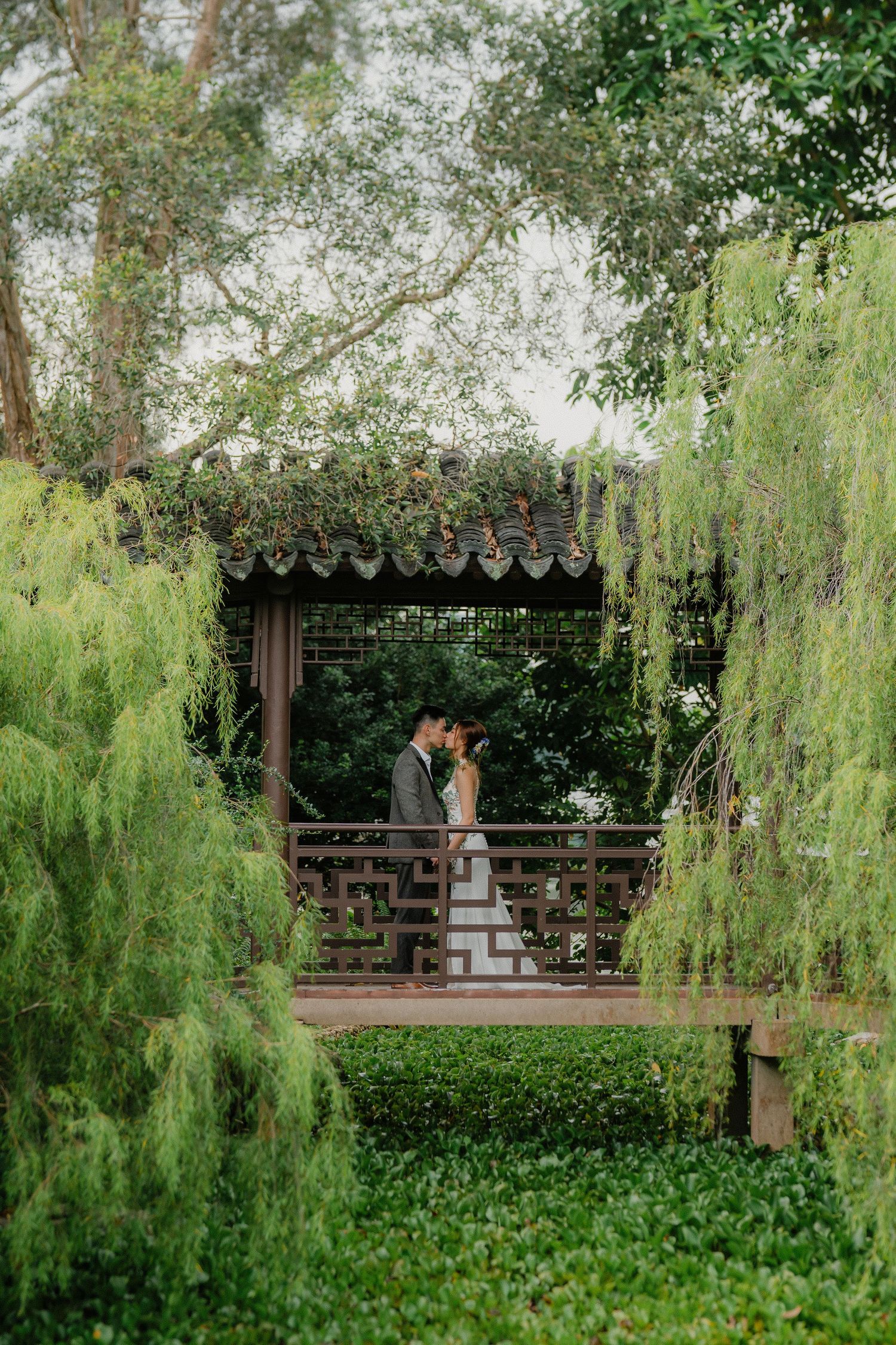 Wooden Chinese garden pagoda with ornate details sits among drooping willow trees and lush greenery.