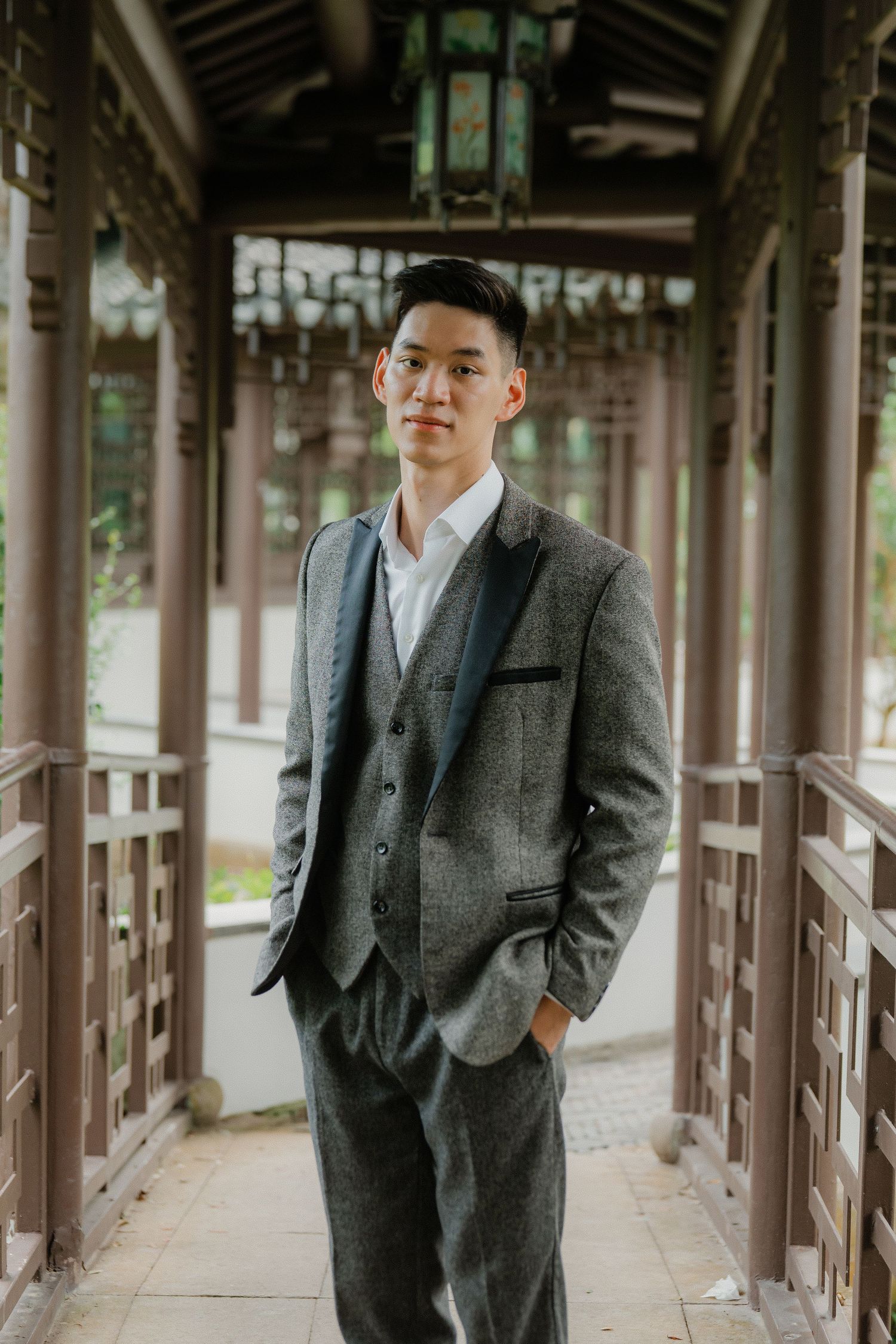 Someone wearing a grey tweed suit and flat cap stands in a covered outdoor wooden corridor walkway.