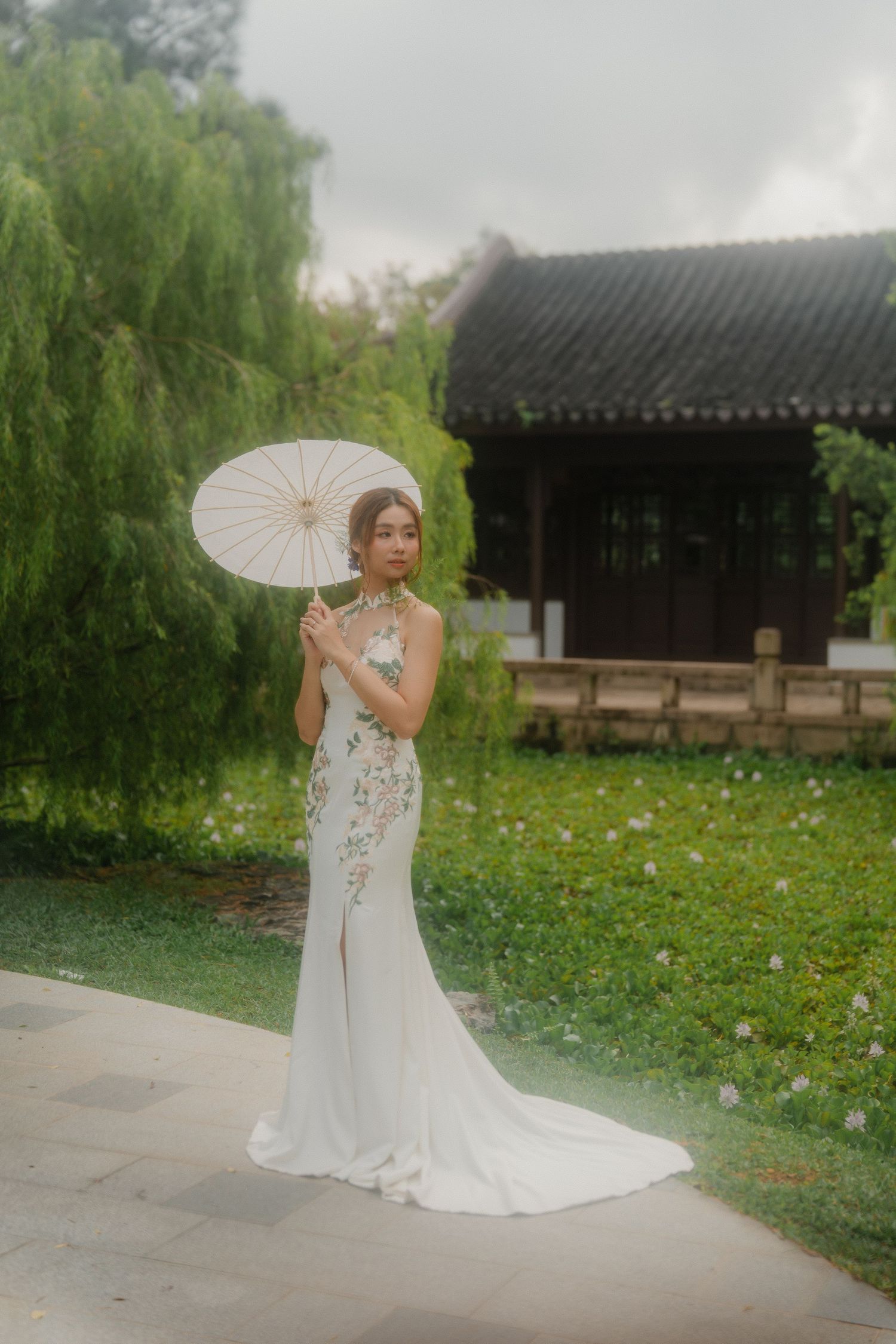 A romantic bridal portrait in a garden setting with a white parasol and flowing wedding gown against traditional architecture.