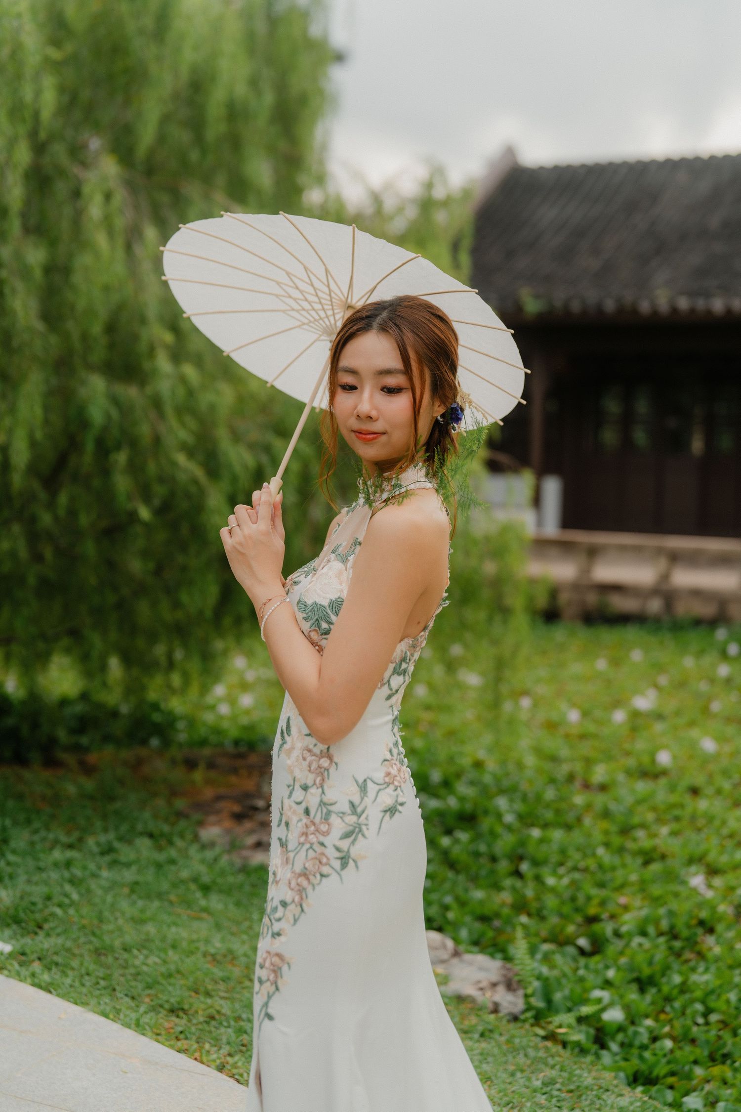 A bride in a white floral dress poses with a traditional paper parasol in a Chinese garden setting.