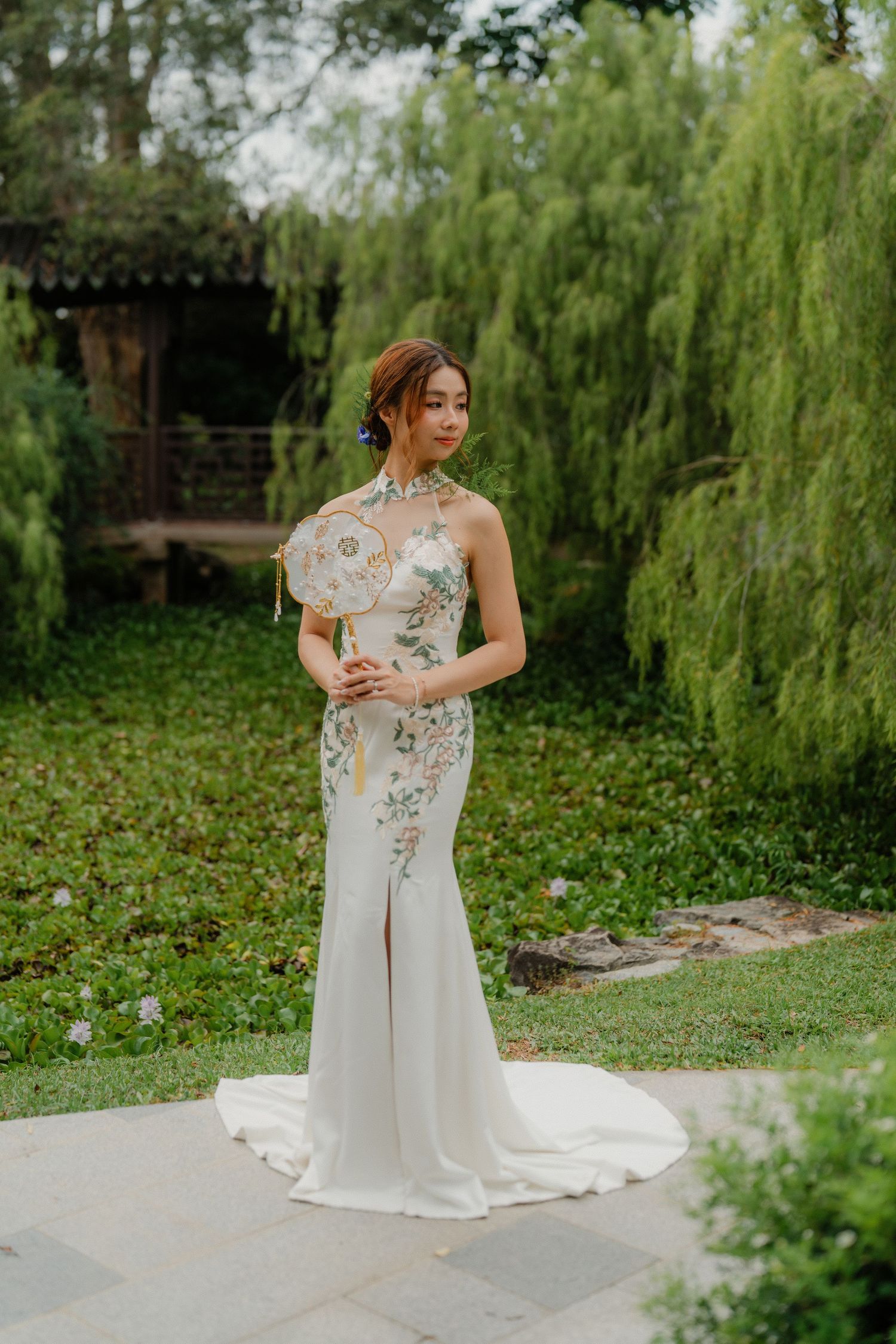A graceful white wedding gown with floral embroidery photographed against weeping willows in a garden.