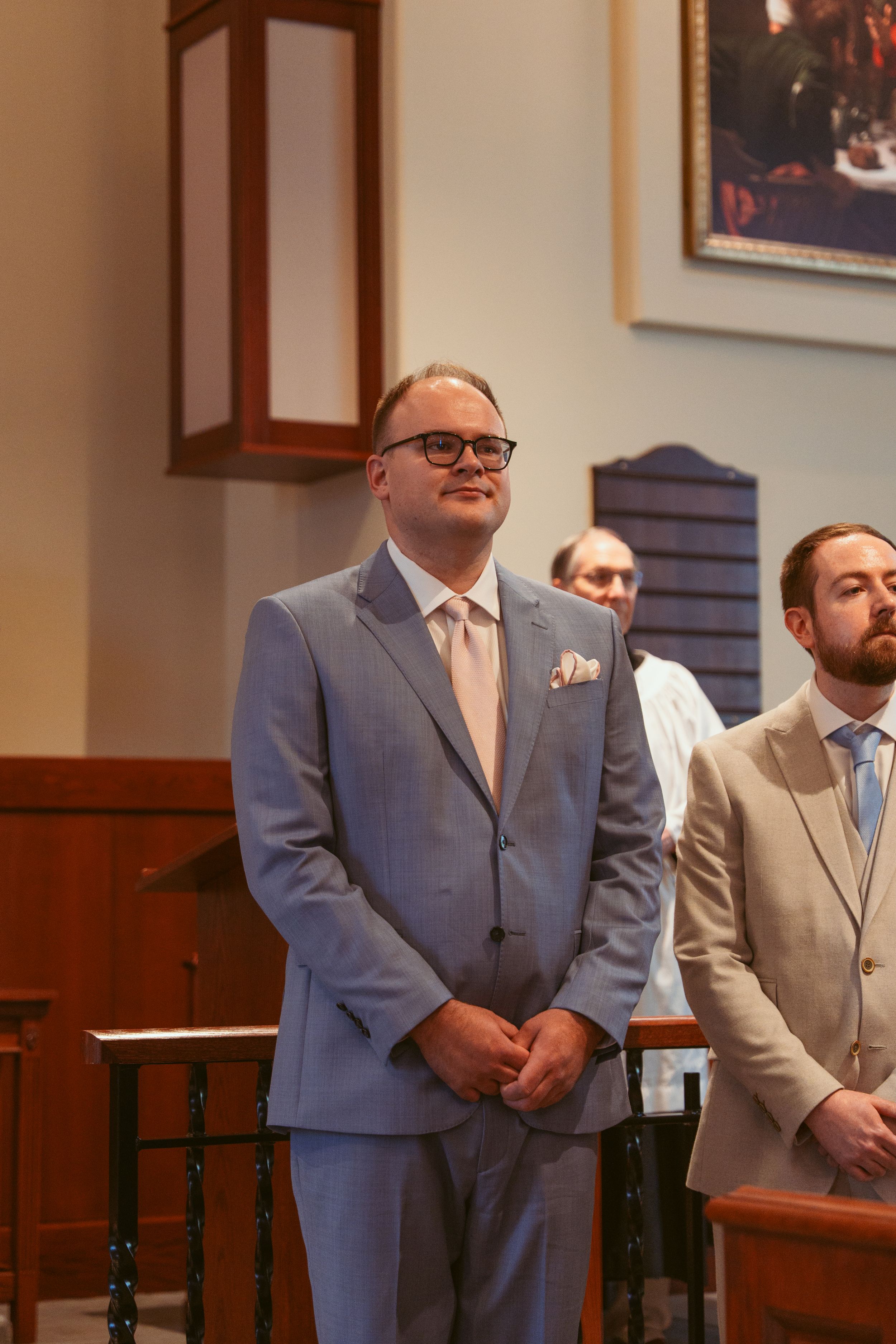 An expectant groom in a light blue suit and glasses stands at the altar during a traditional church wedding ceremony.