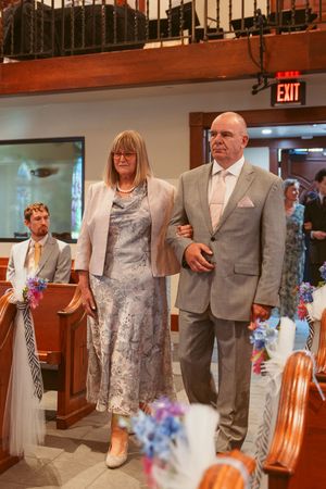 Wedding attendants make their way down the aisle adorned with blue and pink floral decorations in a church setting.