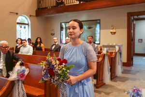A bridesmaid in a light blue dress carries a colorful bouquet during a church wedding procession.