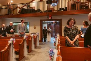Wedding guests gather inside a church sanctuary with wooden pews decorated with bows and flowers.