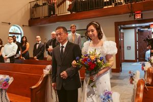 A bride in a white dress walks down the church aisle holding a vibrant bouquet as guests look on from wooden pews.