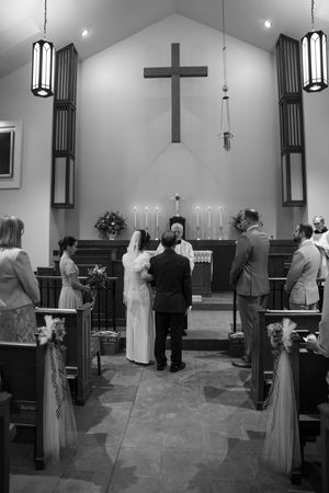 A traditional church wedding ceremony takes place at the altar with hanging pendant lights and a large wooden cross.