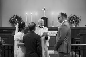 A black and white photograph captures an intimate wedding ceremony with candles and floral arrangements at the altar.