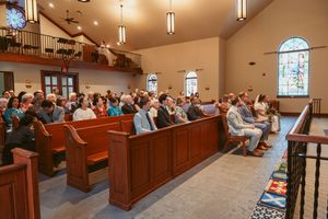 A congregation sits in wooden pews inside a church sanctuary with stained glass windows and pendant lighting.