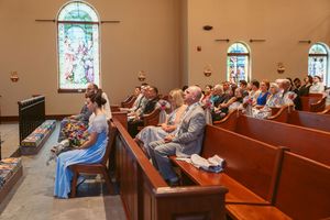 Church congregation sits in wooden pews during a service with stained glass windows visible in the background.