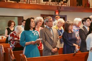 Congregation members stand together during a church service while holding hymnals and singing.