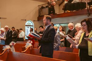 Church congregation members stand in wooden pews decorated with white bows while holding hymnals during service.