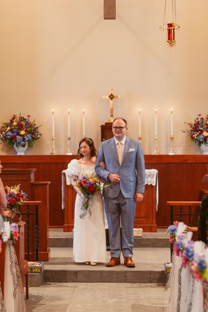 Two people stand at altar in traditional church wedding ceremony with colorful floral arrangements and glowing candles.