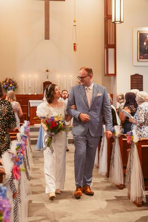 A wedding ceremony recessional in a church sanctuary with floral decorations and candles lining the aisle.