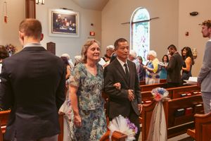 A couple in formal attire walks down a church aisle decorated with white bows as wedding guests look on.