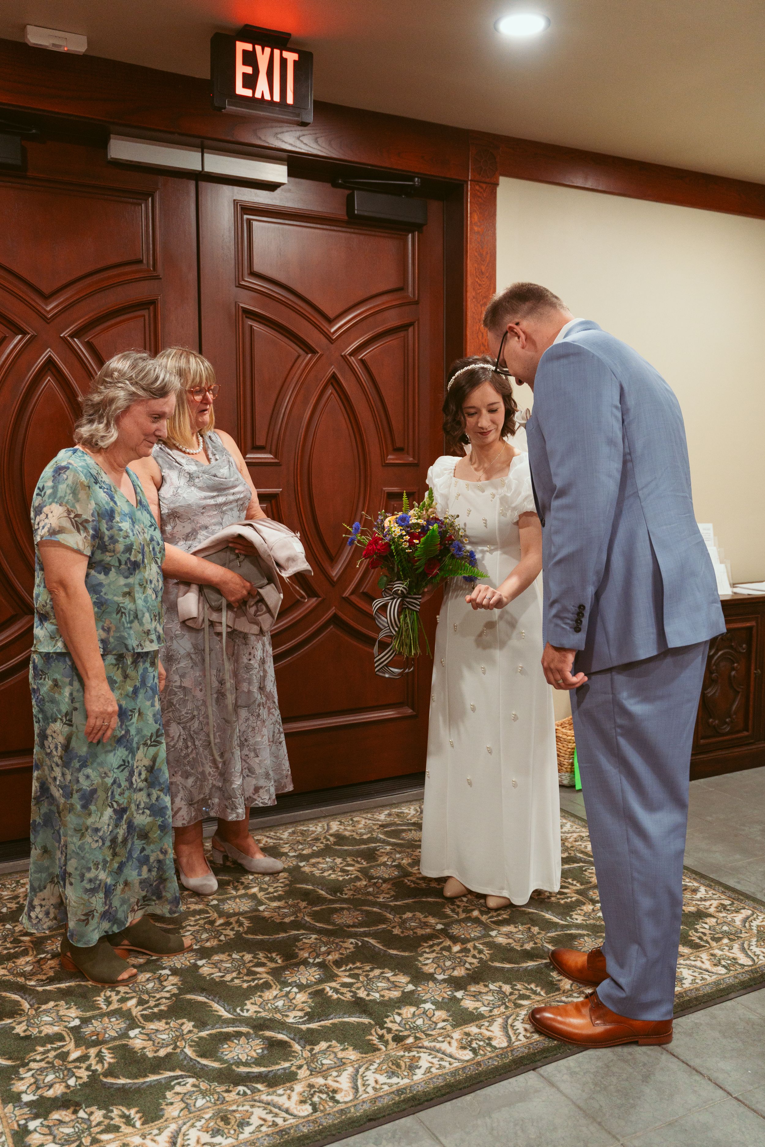 Wedding gathering near ornate wooden doors with floral bouquet and patterned carpet.