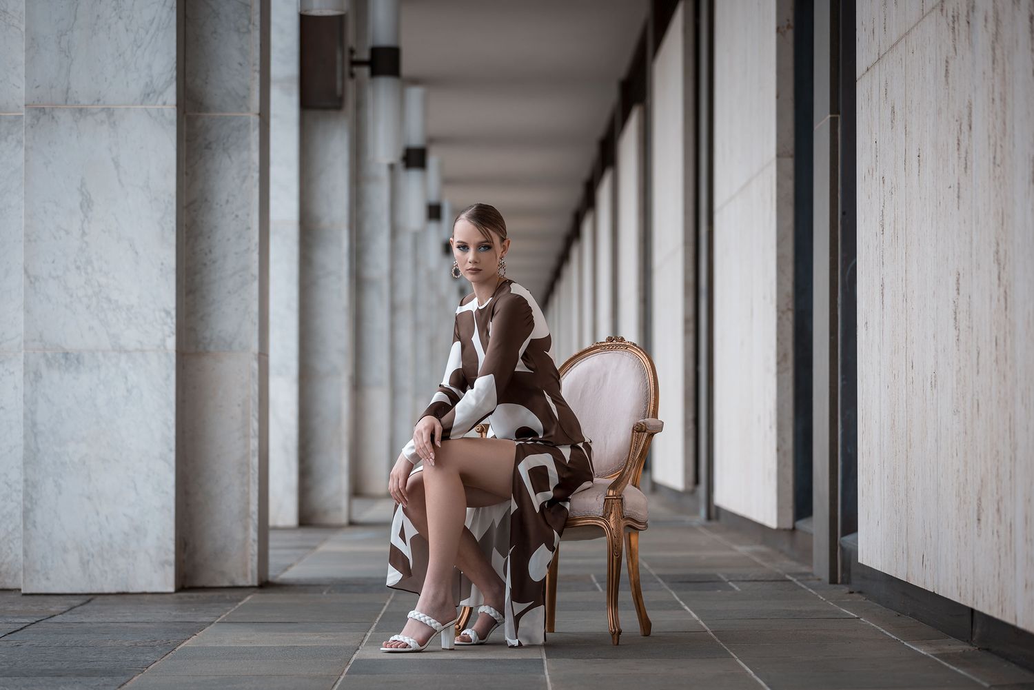 Sophie, Canberra Model sits elegantly in a vintage pink chair along a white columned corridor with dramatic lighting.