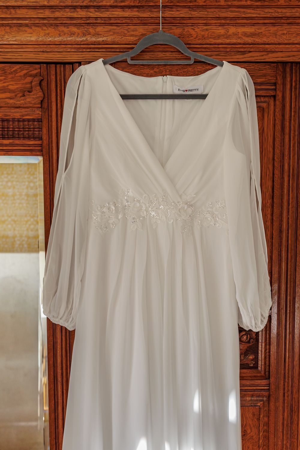 White cotton dress with long sleeves and v-neckline displayed against dark wooden cabinet background.