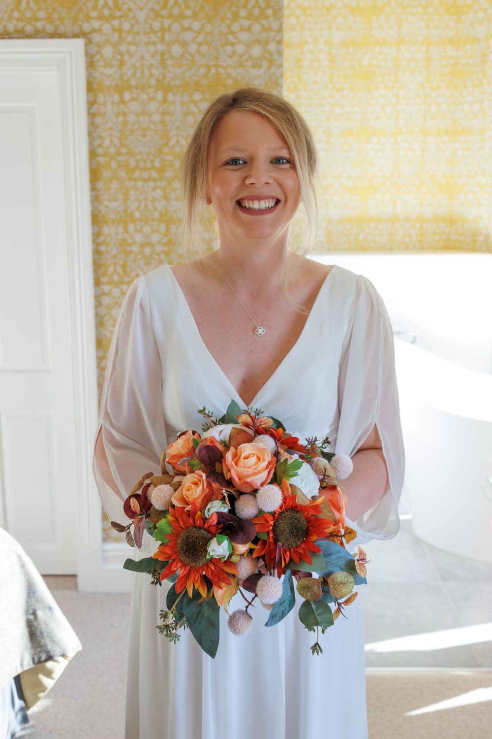 Someone in a white wedding dress holds a vibrant bouquet with orange flowers and greenery against a yellow patterned wall.