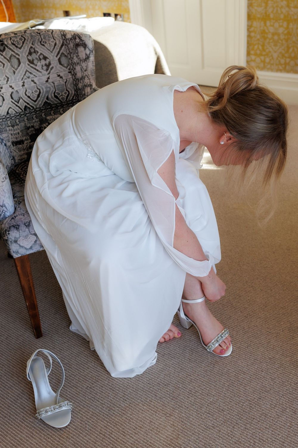 A bride in a white dress adjusts her shoe while sitting in an elegantly decorated room.