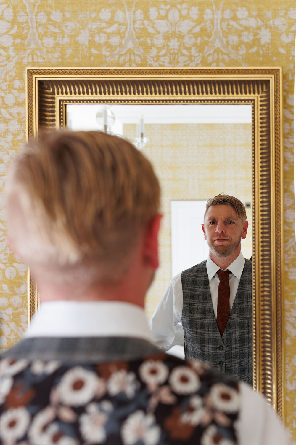 Reflection in ornate gold-framed mirror against yellow damask wallpaper shows wedding preparations.