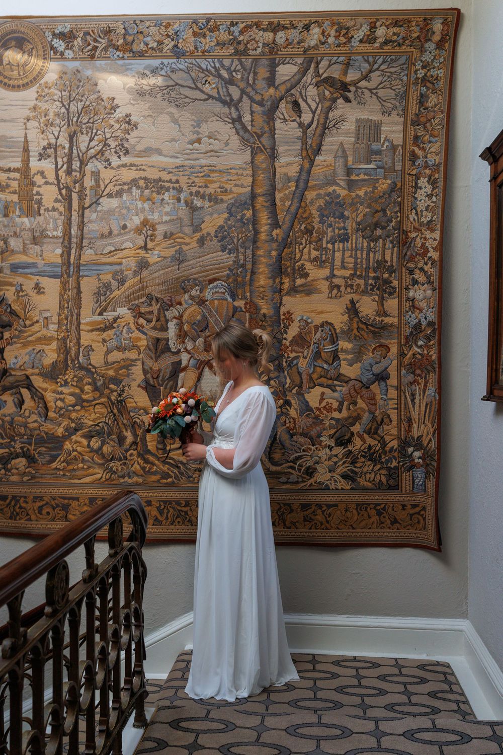 Person in white dress poses near ornate tapestries in an elegant hallway.