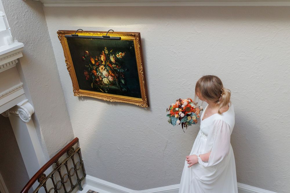 A bride holding a bouquet admires artwork while standing in an elegant stairwell.