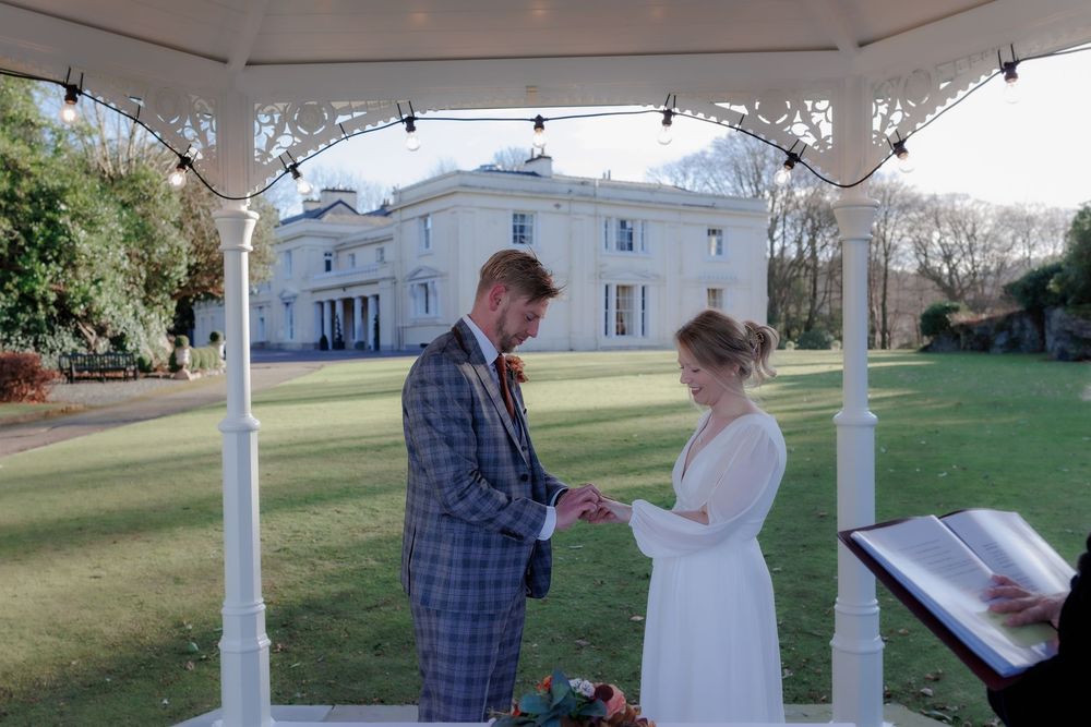 A couple exchanges rings during an outdoor wedding ceremony under a white gazebo with a grand white estate in the backdrop.