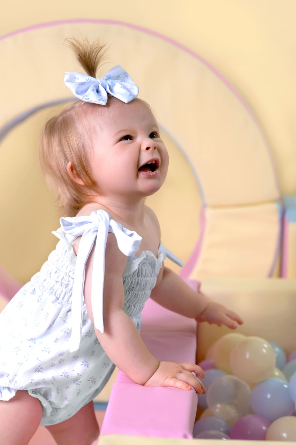 A small toddler wearing white lace plays and looks up joyfully in front of a pastel-colored curved background.