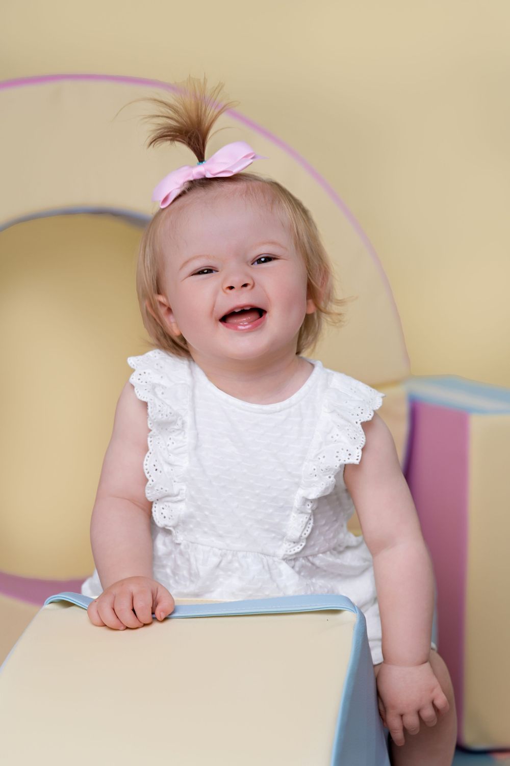Joyful baby in white ruffled dress and pink bow sits on blue play structure.