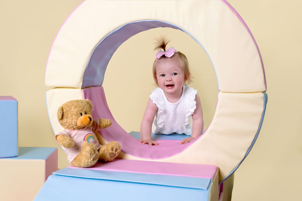 Happy baby plays in curved playroom structure with teddy bear beside them.