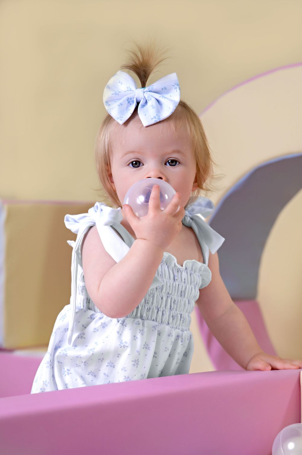 Baby in white dress with blue bow holds toy while playing in pink playroom setting.