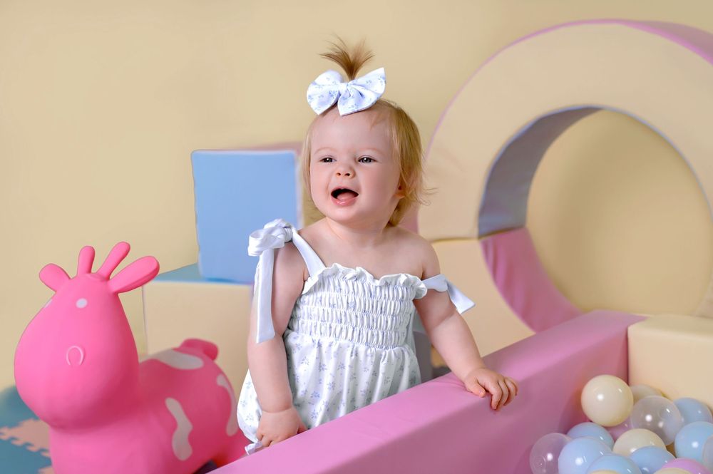 White dress-clad baby laughing near pink rocking horse and decorative moon background.