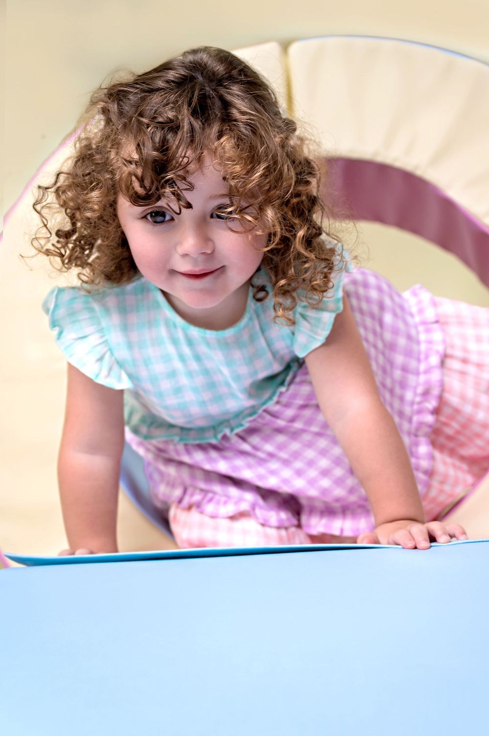 Child wearing gingham dress and white blouse lies down while looking upward with curly hair.