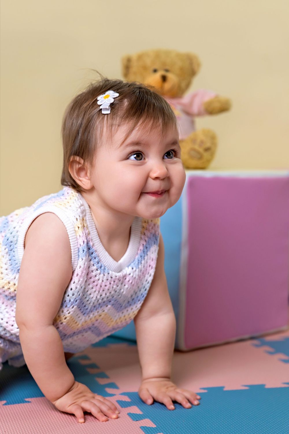A smiling baby in a white polka dot outfit crawls on colorful foam puzzle mats with a teddy bear in the background.