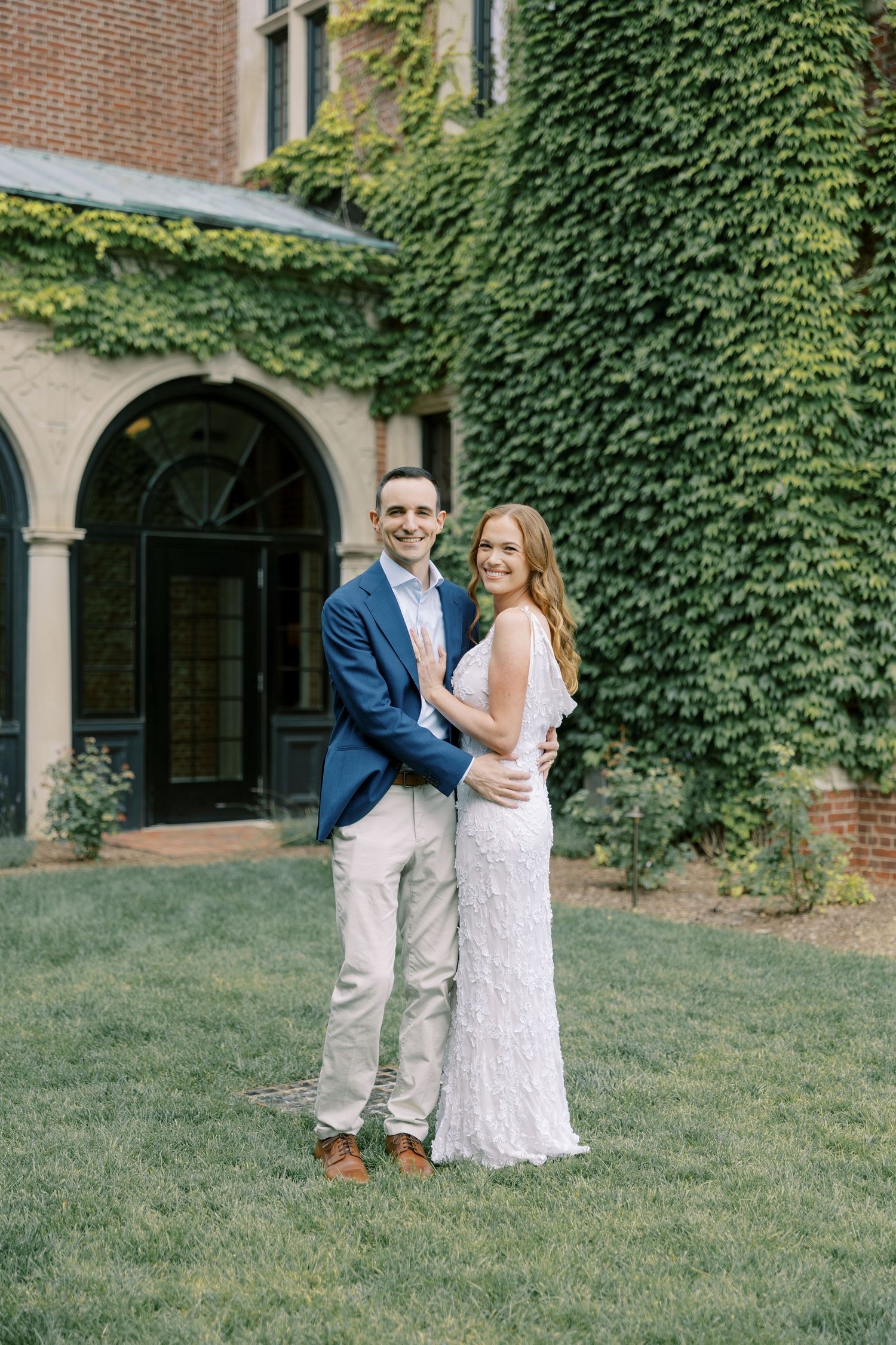 A couple embraces on a grassy lawn in front of an ivy-covered brick building during their special day at Pendry Natirar in Peapack, NJ.