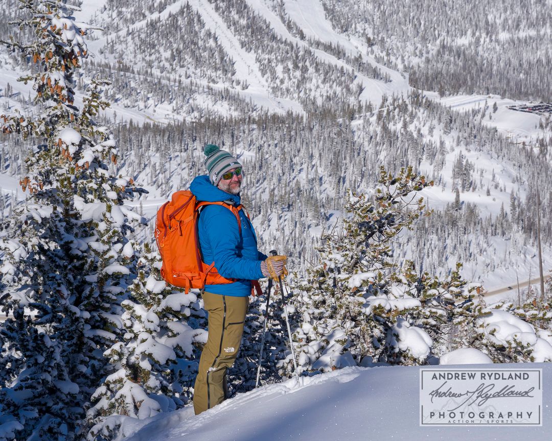 Winter hiker with orange backpack overlooks snow-covered mountain valley and ski slopes.