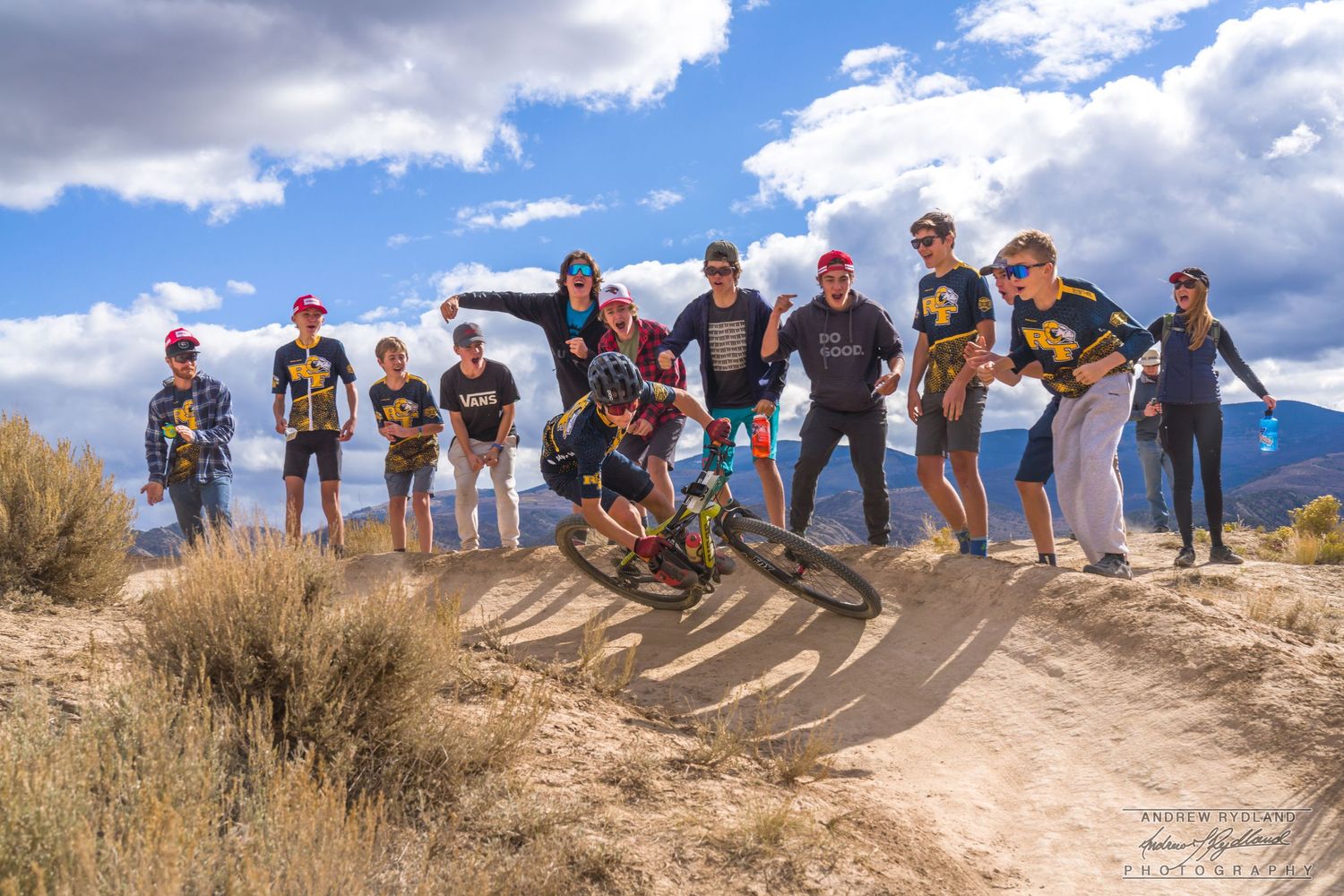 Group of mountain bikers gathered on dirt jump trail against mountain landscape.