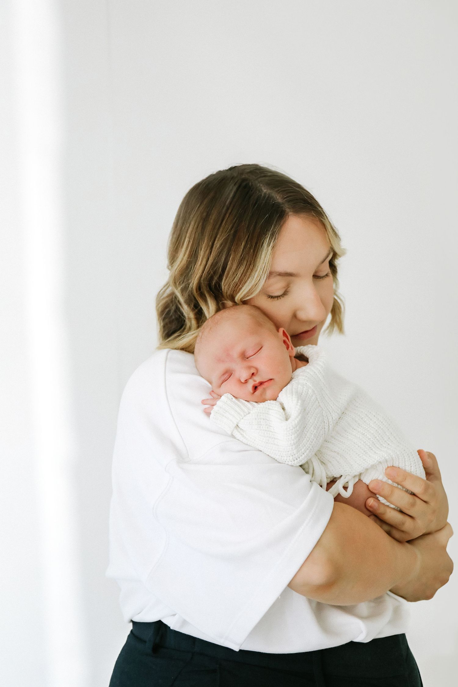 A tender moment shows a mum cradling a sleeping newborn baby wrapped in white against a light background, during newborn shoot in Nottingham