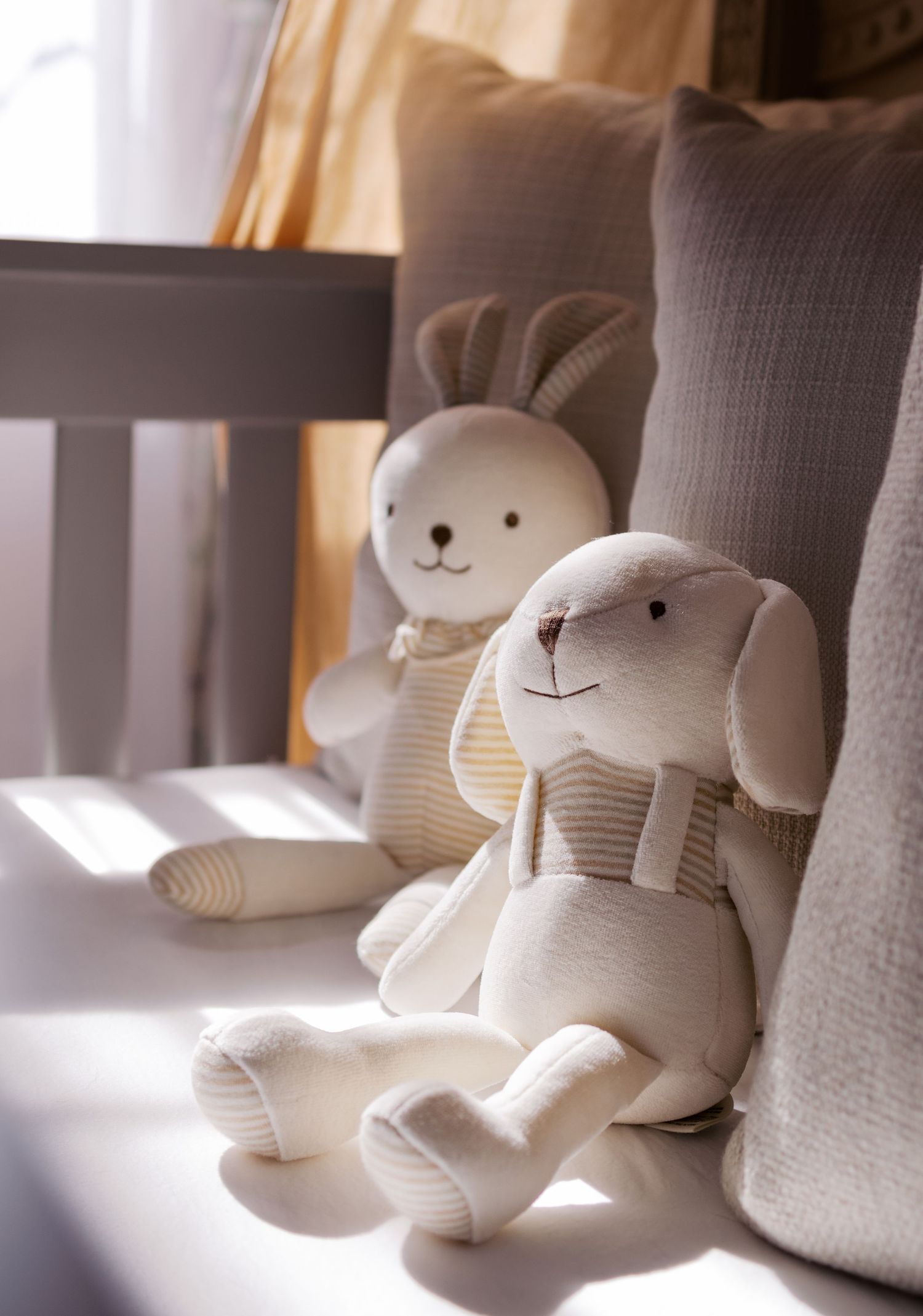 Two plush white bunny toys sitting on a gray couch in soft window light.
