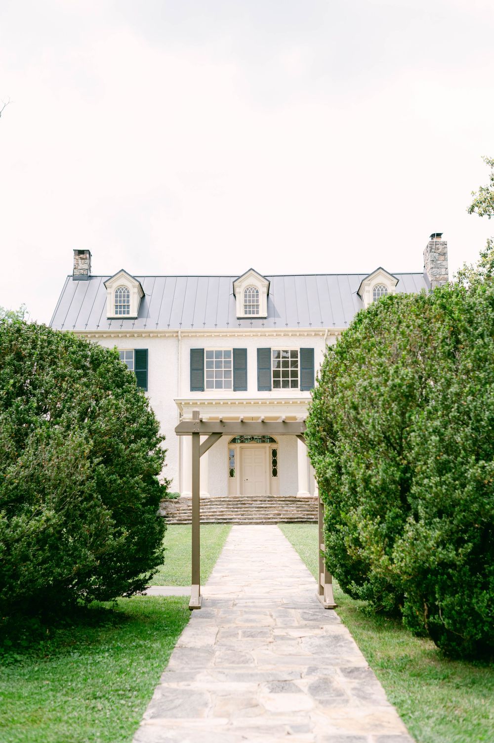 A white farmhouse at Rust Manor House with dormers and a pergola is connected by a concrete walkway through manicured shrubs and lawn.