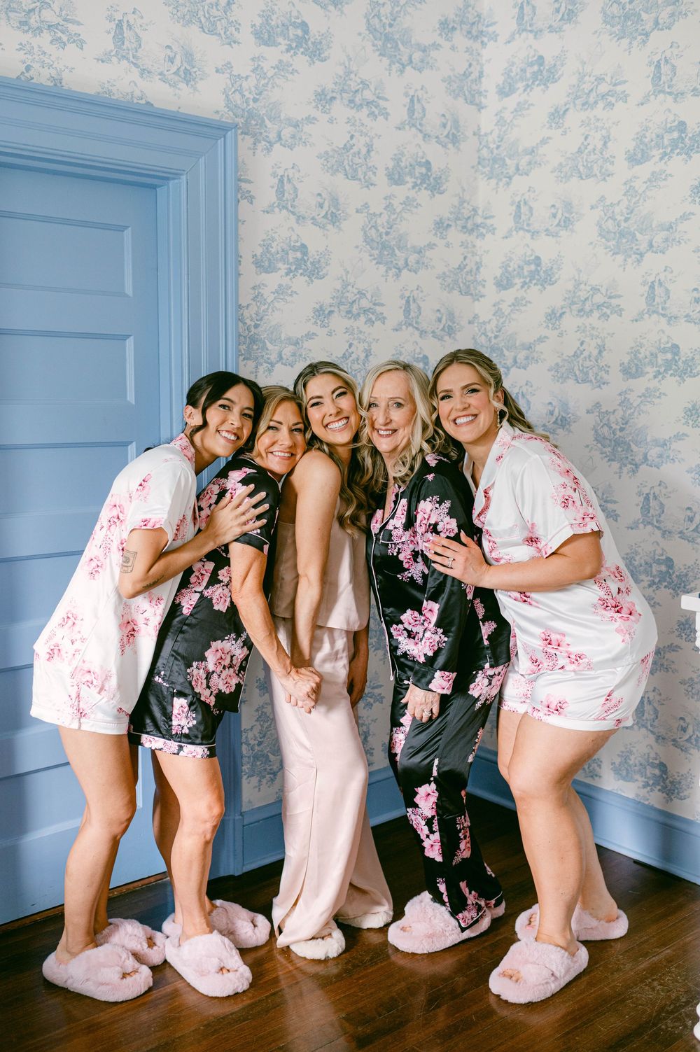 Group of friends in matching floral sleepwear and fuzzy slippers pose together against blue toile wallpaper.